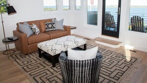 A modern living room with a brown sofa, patterned cushions, a coffee table, and a patterned rug. Large windows offer a view of the sea and a dock with chairs.