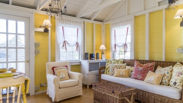 Una habitación acogedora con suelo de madera, un sillón blanco, un sofá con cojines estampados y una cesta de mimbre.