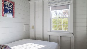 A room with white paneled walls, a single bed with a white bedspread, a window with a view of trees, and a wall-mounted light switch. - Sweet clapboard cottage that is walk to the beach and downtown! (Chatham)