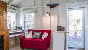 A cozy room with a red sofa, wooden cabinets, and a window overlooking a balcony. - Sweet clapboard cottage that is walk to the beach and downtown! (Chatham)