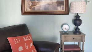 Ein Zimmer mit einem gerahmten Strandbild, einem Schild mit der Aufschrift 'BEACH HOUSE', einem Ledersofa mit rotem Kissen, einem kleinen Holfnachtkästchen mit Lampe und einer Uhr.