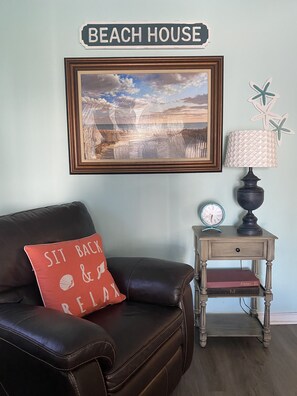 A room with a framed beach scene, a 'BEACH HOUSE' sign, a leather couch with a red pillow, a small wooden nightstand with a lamp, and a clock.