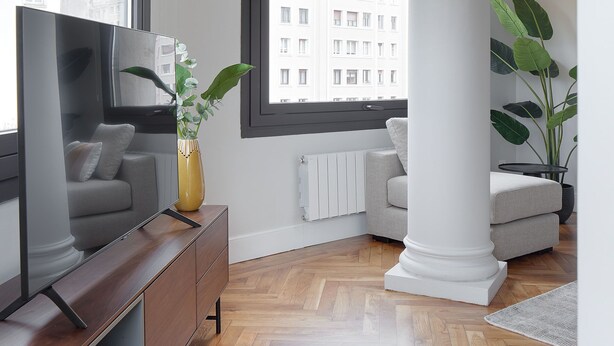 Uma sala de estar moderna com piso de madeira, uma TV de tela plana grande, um sofá, uma mesa de centro e um vaso com planta.