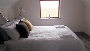A bedroom with a bed, pillows, a bedside lamp, and a window with a view of the beach. - Stunning Tory Island Apartment (Tory Island)