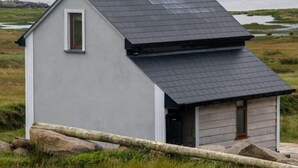 A small, single-story cabin with a dark roof, a window, and a door, situated in a grassy area with a body of water in the background. - Stunning Tory Island Apartment (Tory Island)