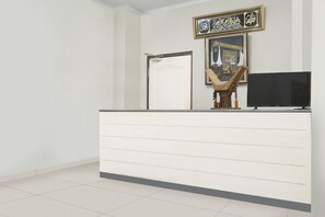 A reception area with a white reception desk, a flat-screen TV, a framed Quran, and a staircase. - OYO 2976 Graha Balfas (Samarinda)