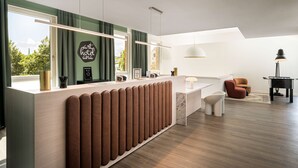 A modern hotel reception area with a large desk, a mirror, and a view of greenery outside. - Arthotel ANA Amber (Rostock)