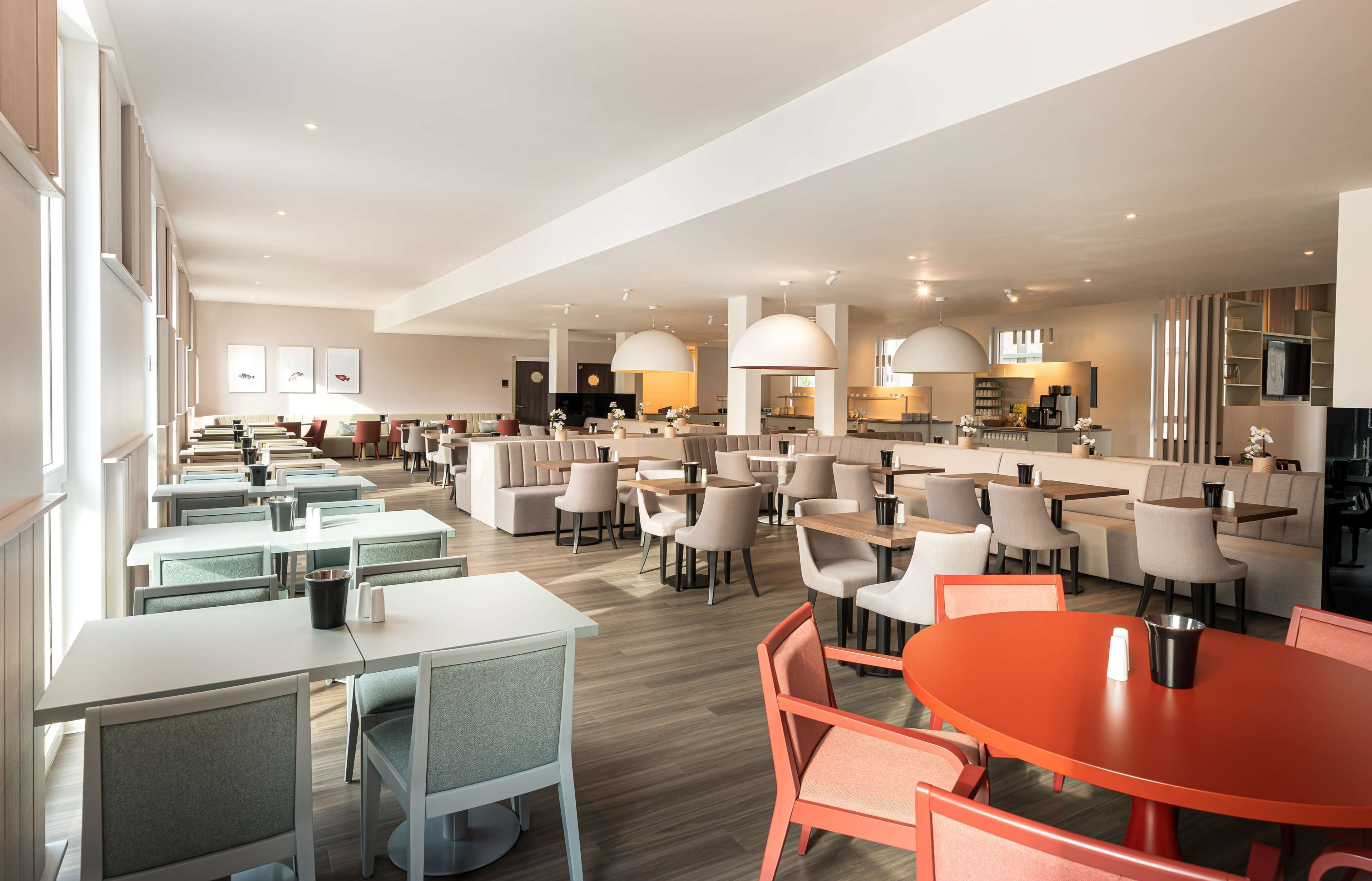 Un'area pranzo moderna con diversi tavoli e sedie, una cucina sullo sfondo e luci a sospensione.