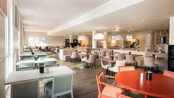 A modern dining area with multiple tables and chairs, a kitchen in the background, and pendant lighting.