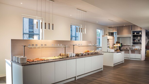 A modern hotel kitchen with a long counter, multiple serving stations, and a clean, minimalist design. - Arthotel ANA Amber (Rostock)