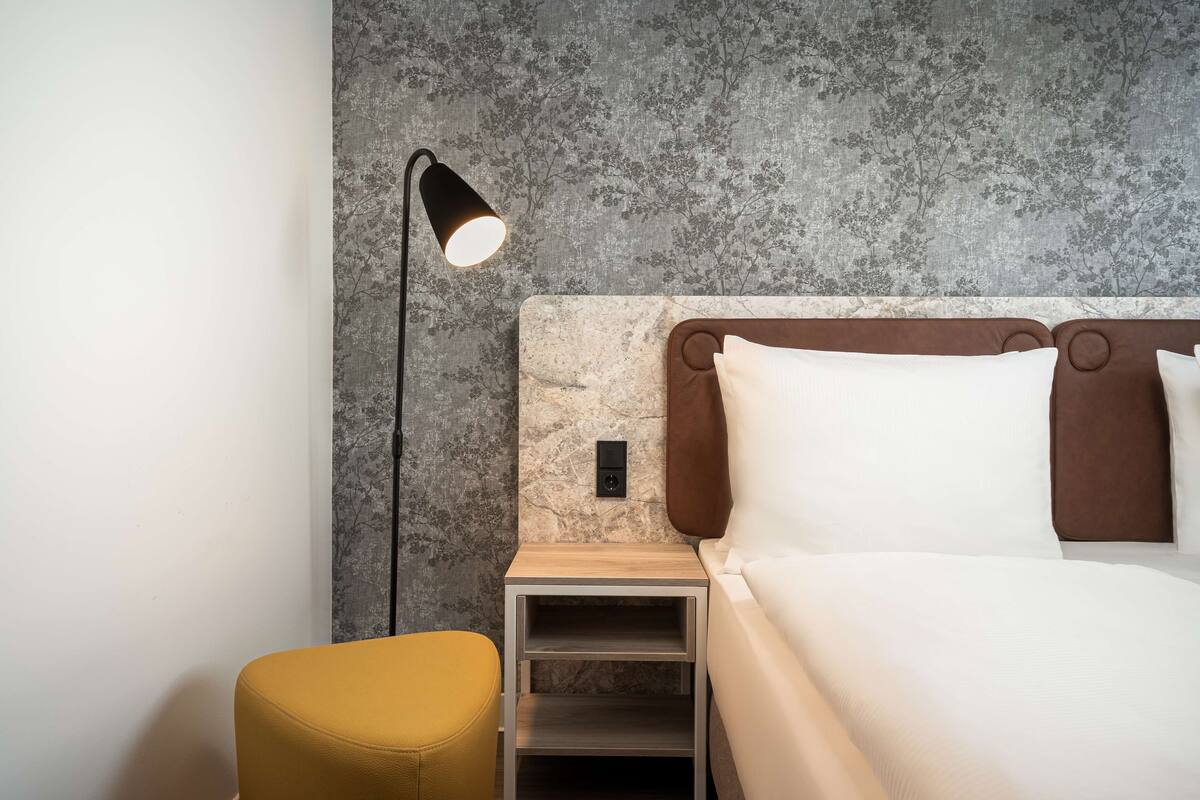 A modern bedroom with a bed, bedside table, lamp, and a small stool.