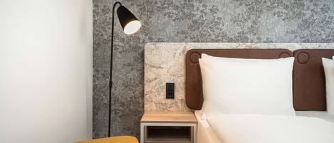 Un dormitorio moderno con cama, mesita de noche, lámpara y un pequeño taburete.