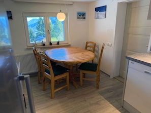 Un comedor con una mesa redonda de madera y cuatro sillas, una ventana con vistas a follaje, y una cocina con electrodomésticos blancos.