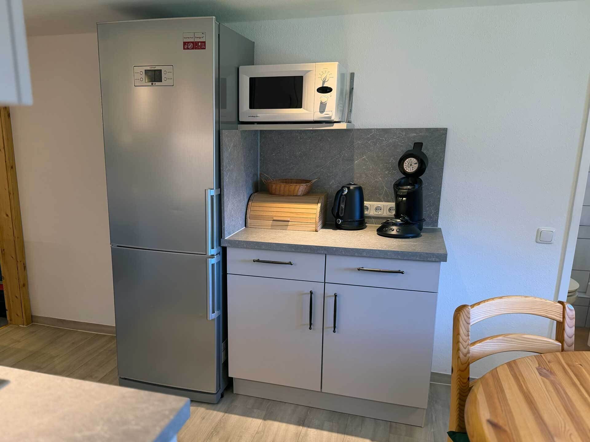 A compact kitchen with a stainless steel refrigerator, a microwave, a coffee maker, and a wooden chair.