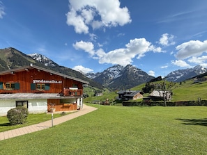 Un bâtiment en bois, sur lequel on peut lire l’inscription « gondamika.at », situé dans une région montagneuse, avec des champs verdoyants et des sommets enneigés en arrière-plan.