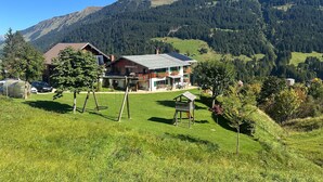 Un chalet de dos pisos con techo inclinado, una terraza de madera y un columpio en un exuberante valle verde con montañas al fondo.