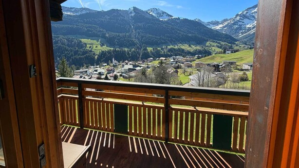 Un balcón de madera con vistas a un pueblo de montaña y cumbres nevadas.