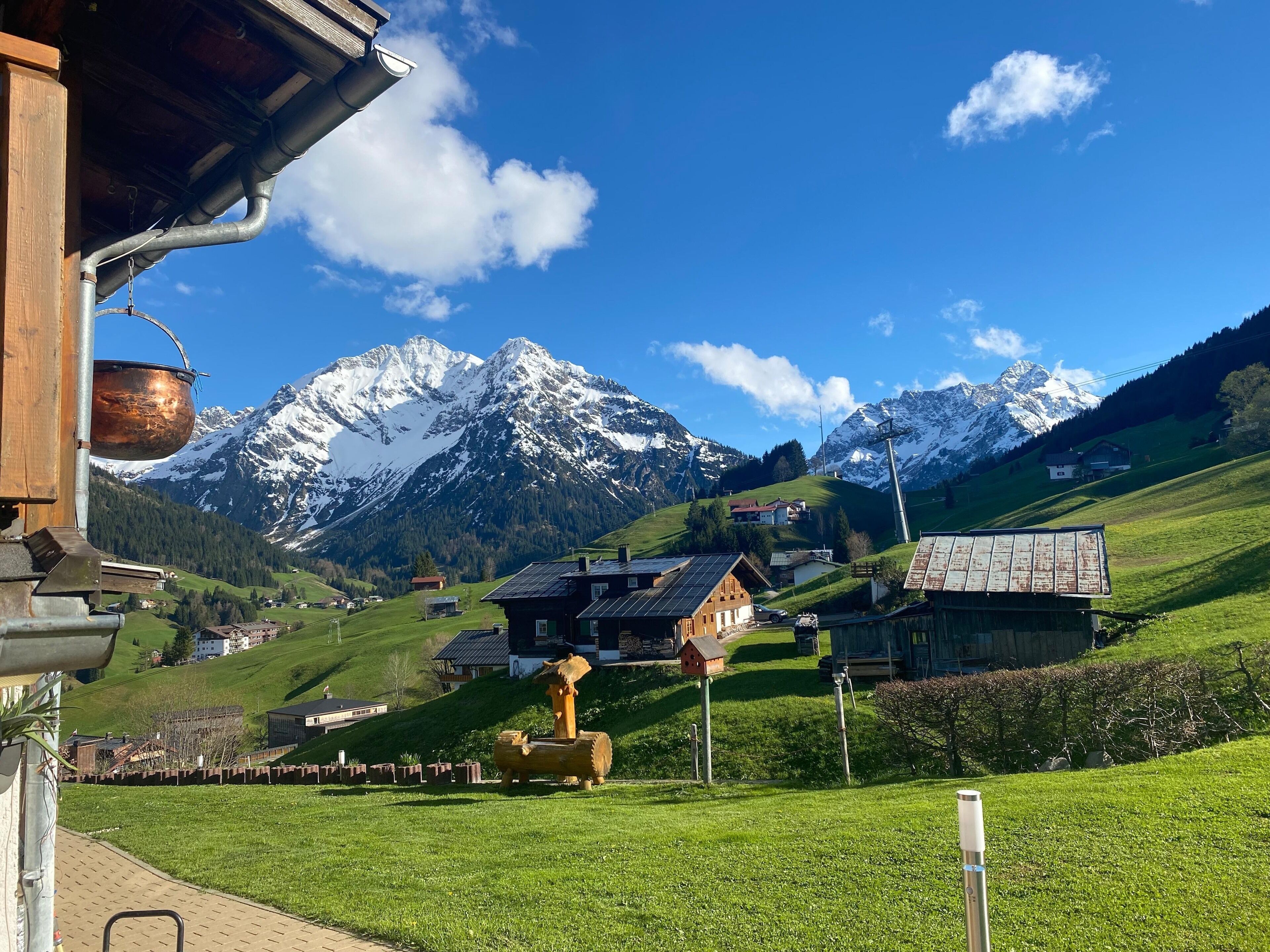 Un pueblo rural con casas tradicionales, una estructura de madera con una olla de cobre y montañas nevadas al fondo.