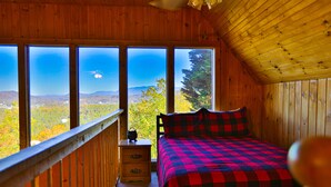 Quarto de cabana de madeira com cama, cômoda e grandes janelas com vista para uma floresta e montanhas.