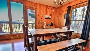 Uma área de refeições com mesa grande e bancos de madeira, um canto para café e grandes janelas com vista para o exterior.