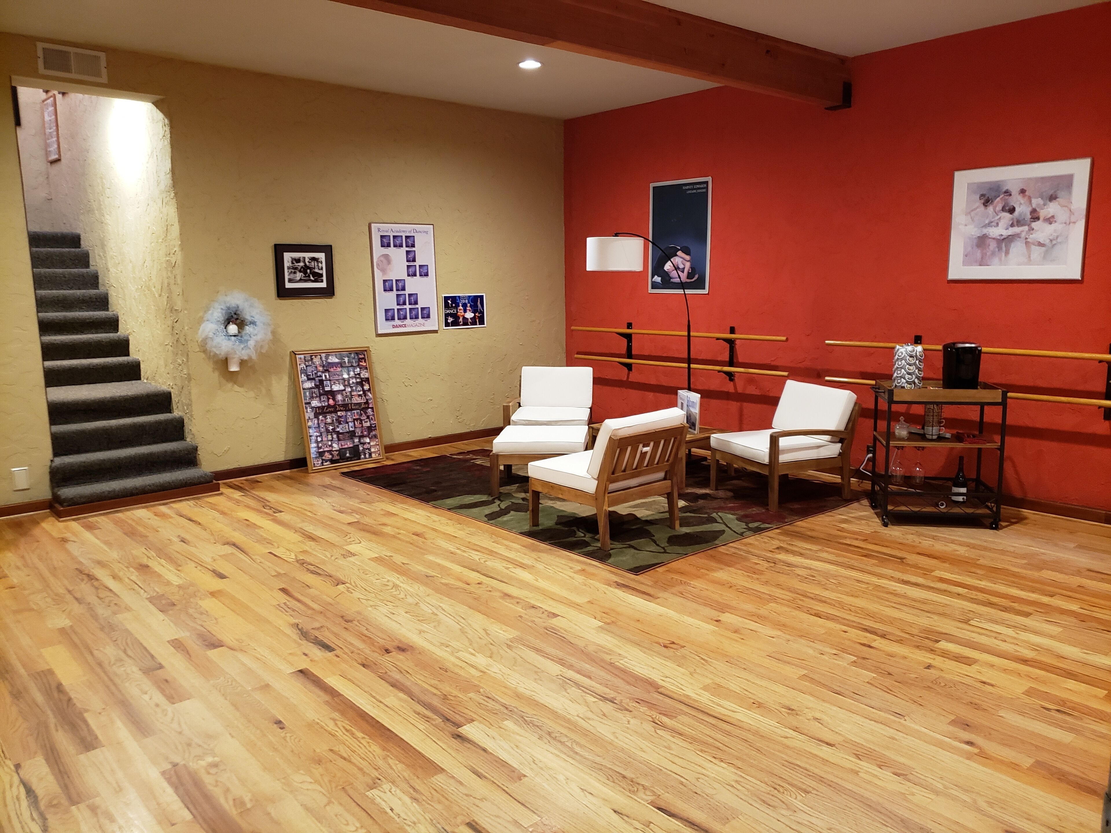Quarto com piso de madeira, escada, cadeiras brancas, uma pequena mesa e uma parede vermelha com cartazes de dança.