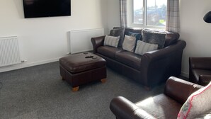 A living room with a brown leather sofa, a matching ottoman, a flat-screen TV mounted on the wall, and a window with curtains.
