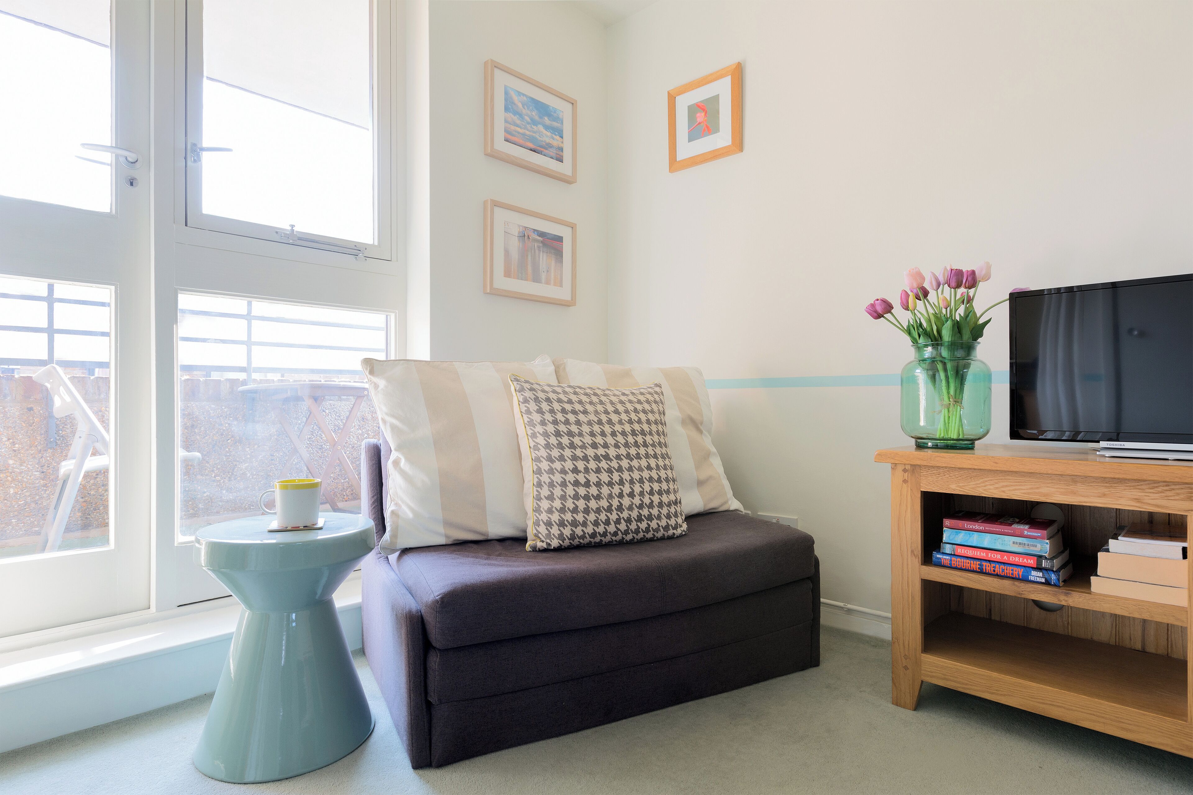 A cozy room with a sofa, a small side table, a TV stand with books, and a window with a view.