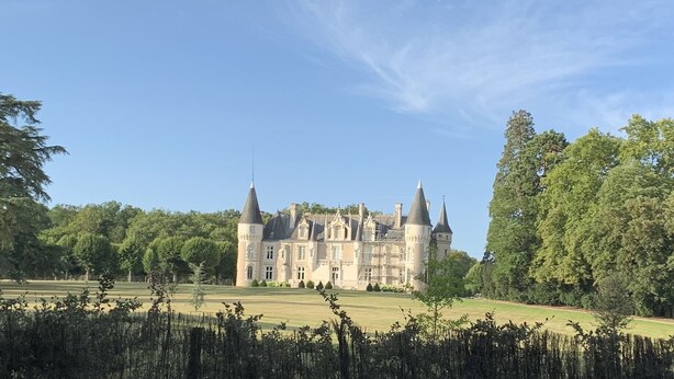 A large, historic building with multiple towers and spires, surrounded by lush greenery and a well-maintained lawn.