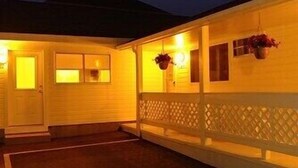 A single-story building with a white exterior, a covered porch with a lattice railing, and hanging flower baskets. - Seashell Cottages/JK Rentals -  The Daniel Unit (Old Orchard Beach)