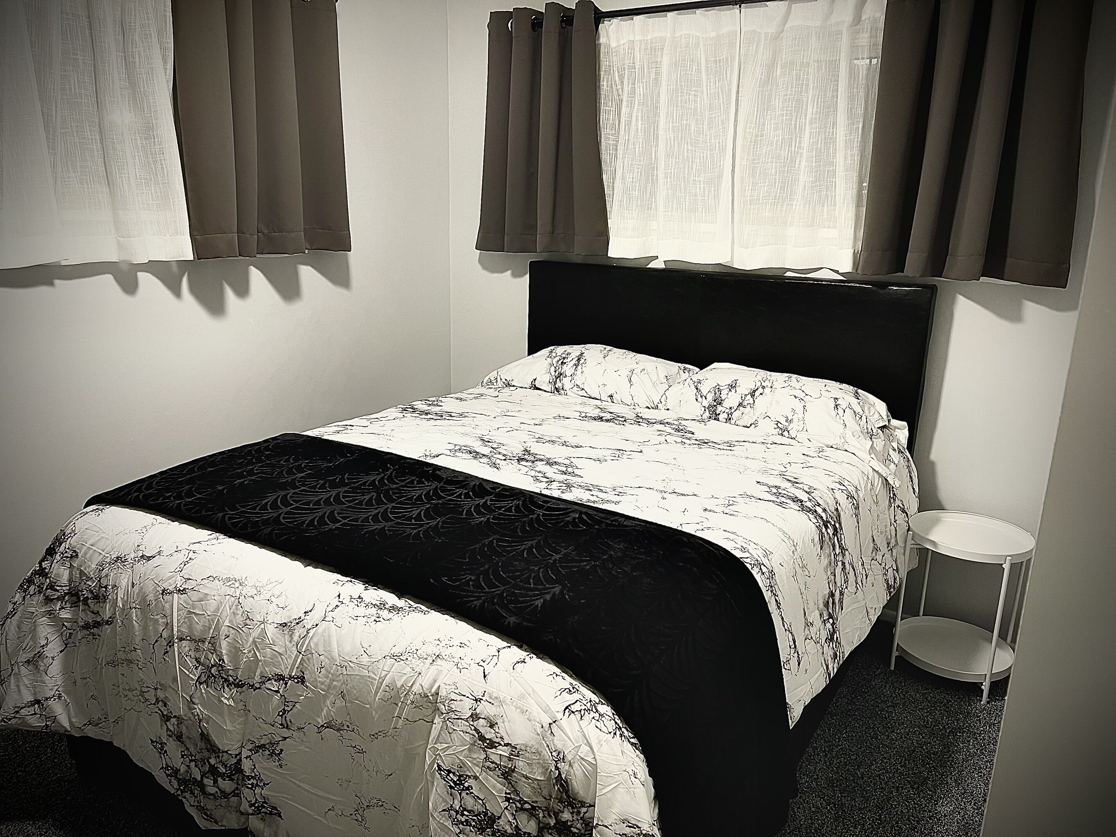 Una cama con una colcha estampada, una mesita de noche y cortinas en las ventanas.