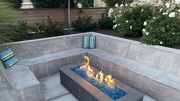Una chimenea rectangular con piedras de vidrio azules y llamas, rodeada por una zona de estar de hormigón con cojines.