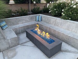 A rectangular fire pit with blue glass stones and flames, surrounded by a concrete seating area with cushions. - Fabulous Urban Oasis w/hot tub and private backyard! (Salt Lake City)