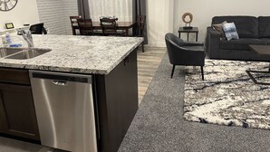 Una cocina moderna con lavavajillas, un comedor con mesa y sillas, una sala de estar con sofá y alfombra, y un reloj de pared.