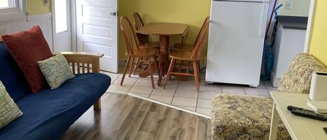 A compact living space with a dining table, chairs, a sofa, a small ottoman, a refrigerator, and a ceiling light fixture.