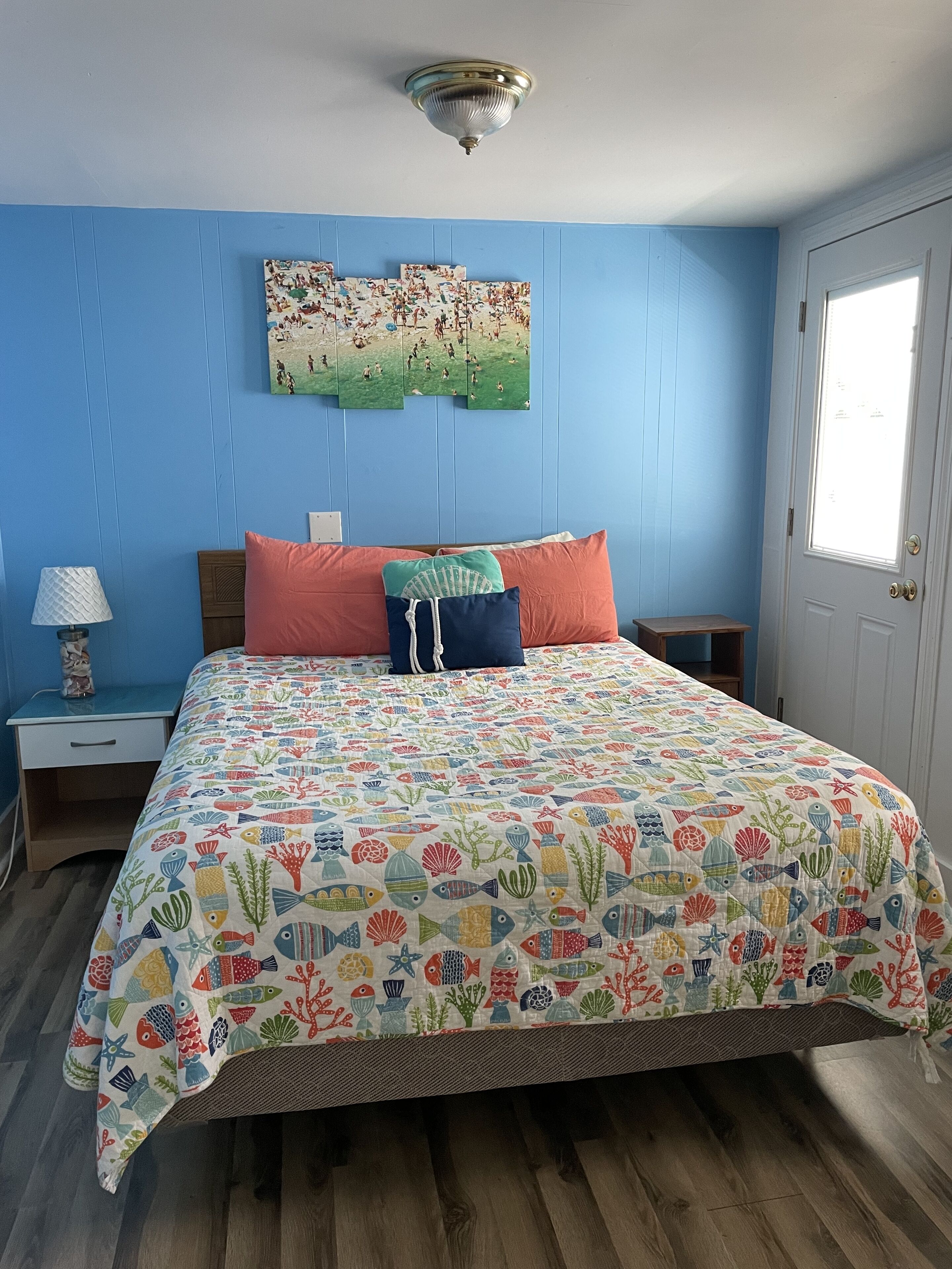 A bedroom with a bed, a nightstand, a lamp, a door, and a wall with a painting.