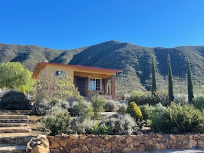 Uma casa térrea com telhado vermelho, rodeada por diversas plantas típicas do deserto e um muro de pedra.