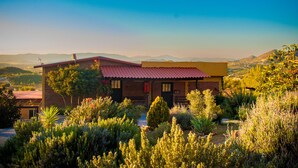 A single-story house with a red-tiled roof, surrounded by lush greenery and a mountainous backdrop.