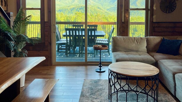 Ein geräumiges Wohnzimmer mit einem großen Holztisch, einem runden Couchtisch und einer Couch. Große Fenster bieten Blick auf eine gebirgige Landschaft.