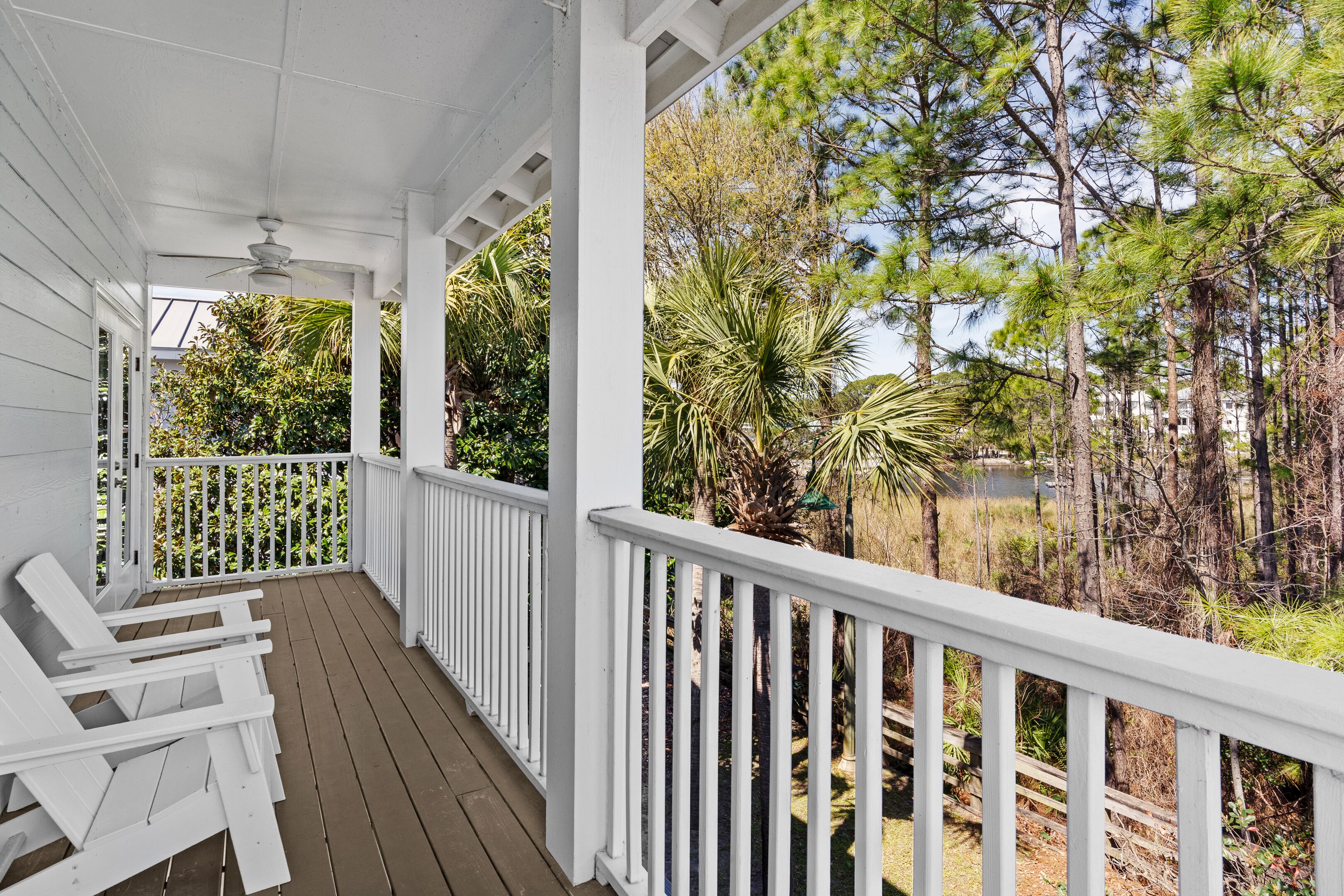 A covered porch with white railings and wooden deck, overlooking a natural landscape with trees and a body of water.