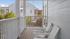 Une véranda couverte, dotée de balustrades blanches et d’un plancher en bois, qui donne sur un quartier résidentiel avec des maisons et des palmiers.
