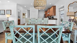 Un coin repas avec une table en bois et des chaises turquoise, un luminaire suspendu et des meubles blancs en arrière-plan.