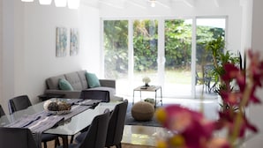 Un espace repas moderne avec une table en verre, des chaises et des luminaires suspendus. On aperçoit un canapé et une petite table avec une plante en arrière-plan.