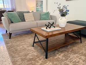 Uma sala de estar com uma mesa de centro de madeira, um sofá com almofadas, um vaso com flores e um quadro na parede.