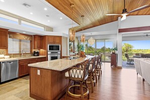 A spacious kitchen with wooden cabinets, a large island, and a dining area with wicker chairs. - LUXURIOUS, REMODELED MAUNA LANI VILLAGES TOWNHOME! PRIVATE BEACH CLUB ACCESS! (Kamuela)