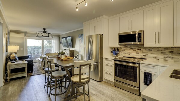 A modern kitchen with white cabinets, stainless steel appliances, a dining area with bar stools, and a view to the outside. - Make Coligny Beach Your Happy Place!! (Hilton Head Island)