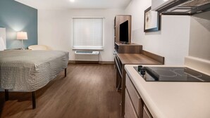 A compact hotel room with a bed, a desk with a built-in stove, a TV mounted on a wooden shelf, and a window with blinds. - WoodSpring Suites Sulphur - Lake Charles (Sulphur)
