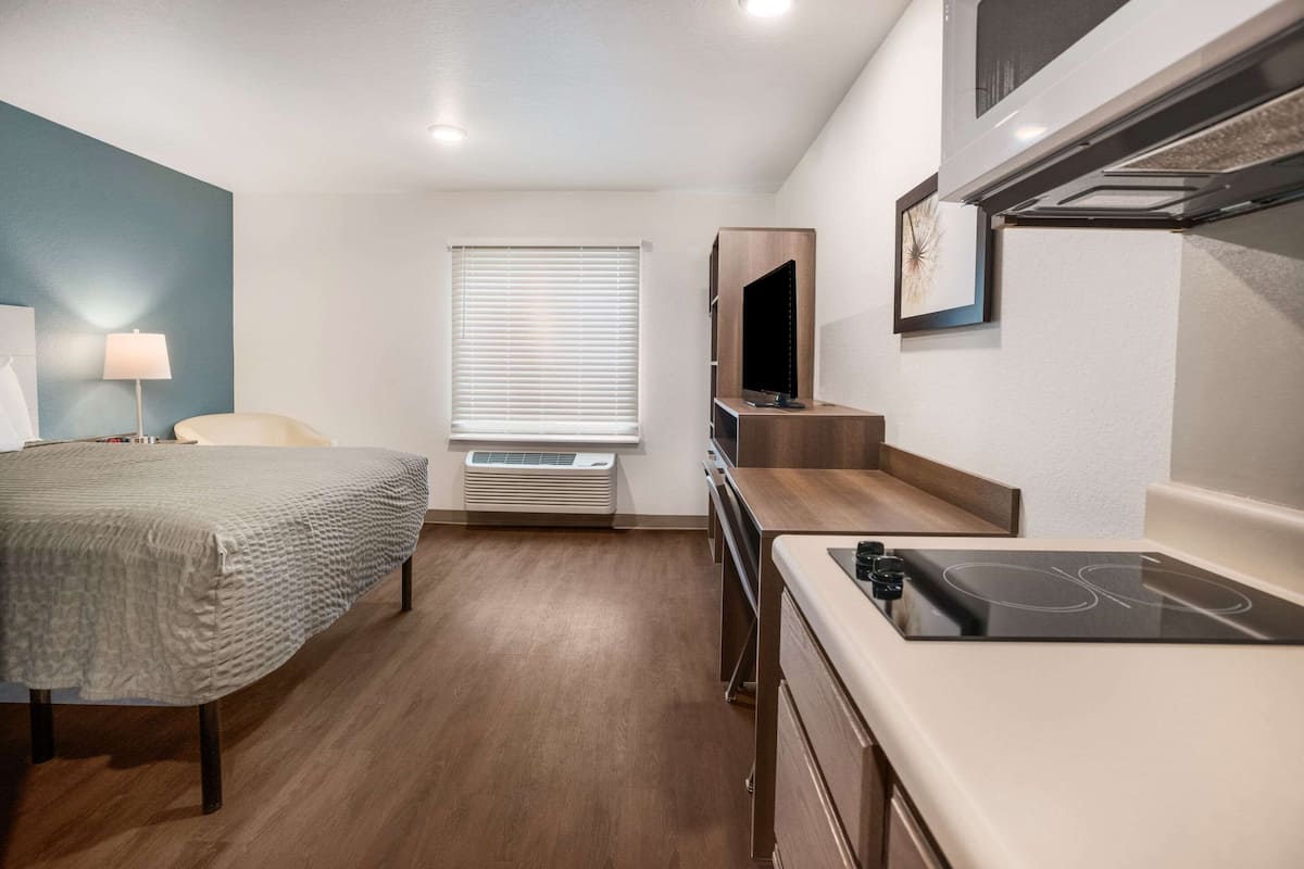 A compact hotel room with a bed, a desk with a built-in stove, a TV mounted on a wooden shelf, and a window with blinds.
