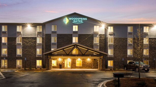 Un hotel de varios pisos con un llamativo letrero de "Woodspring Suites", un acceso cubierto y un área de estacionamiento.