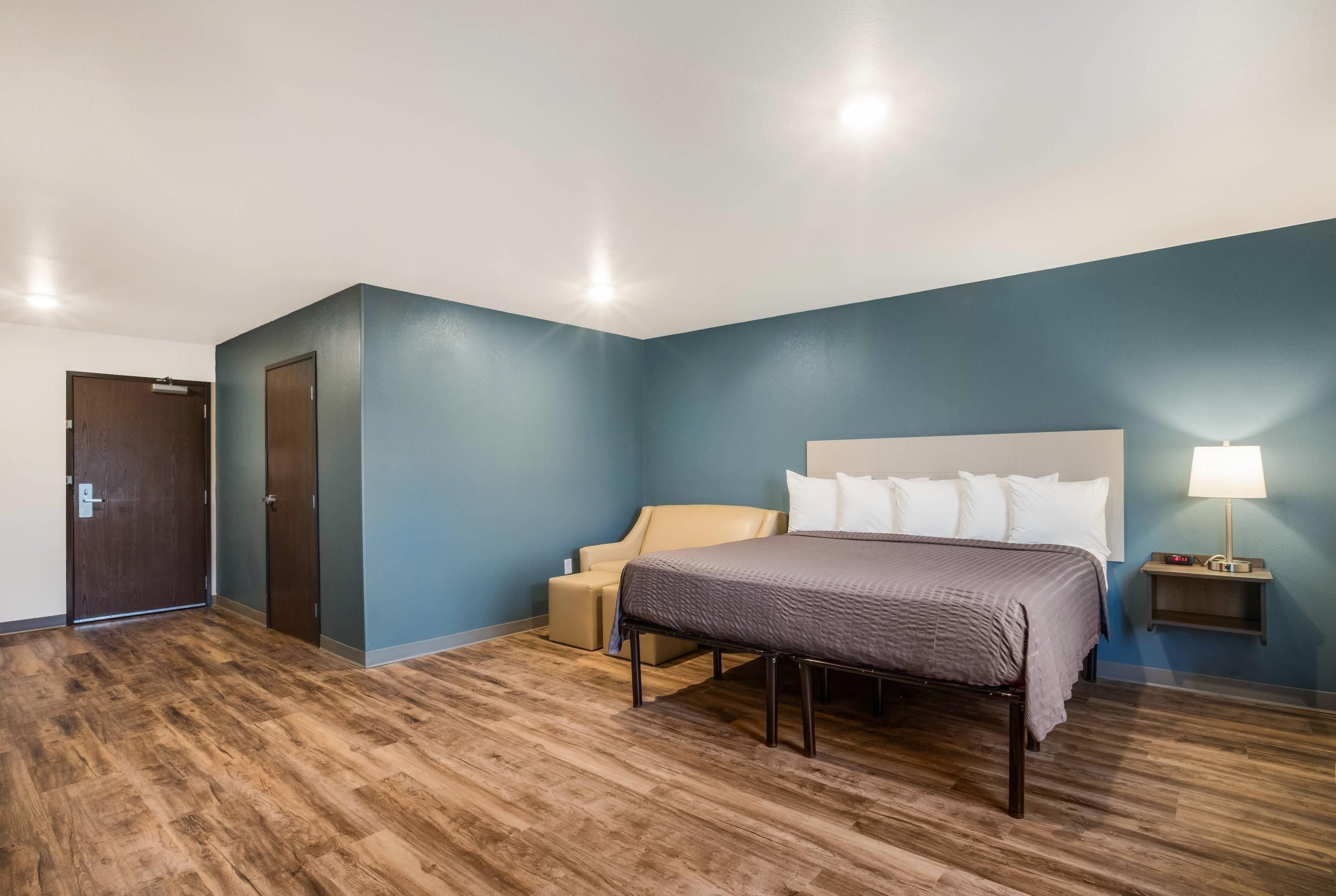 A hotel room with a bed, a chair, a bedside table with a lamp, and a door.
