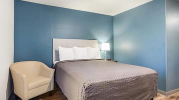 A hotel room with a bed, a chair, a nightstand with a lamp, and a clock.